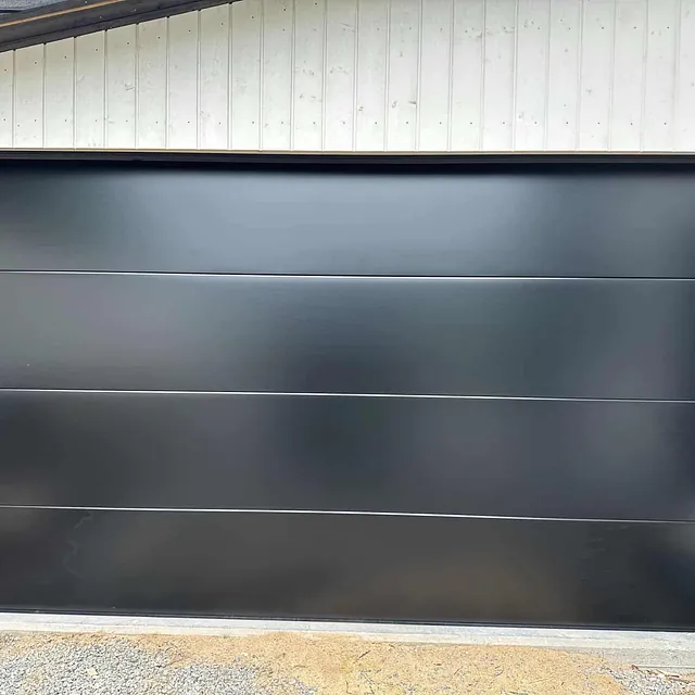 Ny svart garasjeport montert i lys trekledd bygg, sett rett forfra.