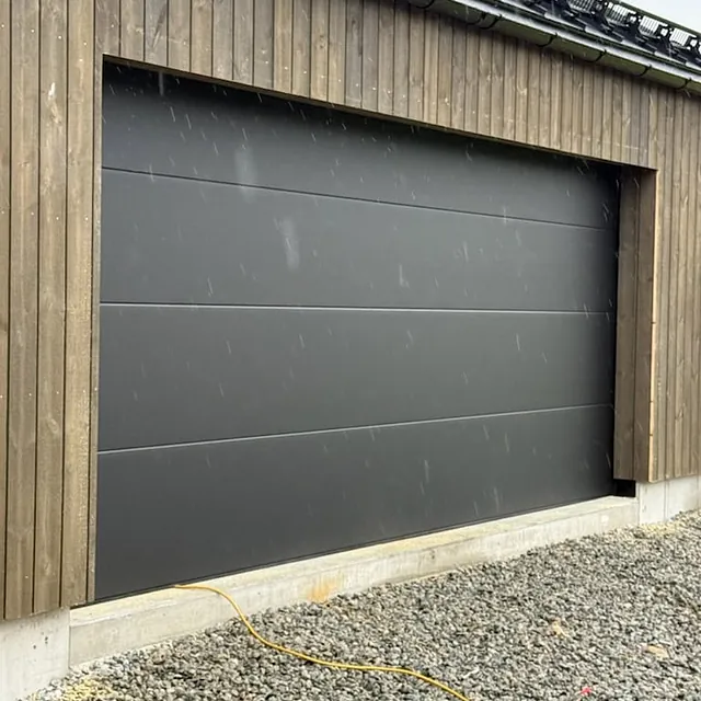 Svart garasjeport montert i nybygg med stående trekledning og grus foran.