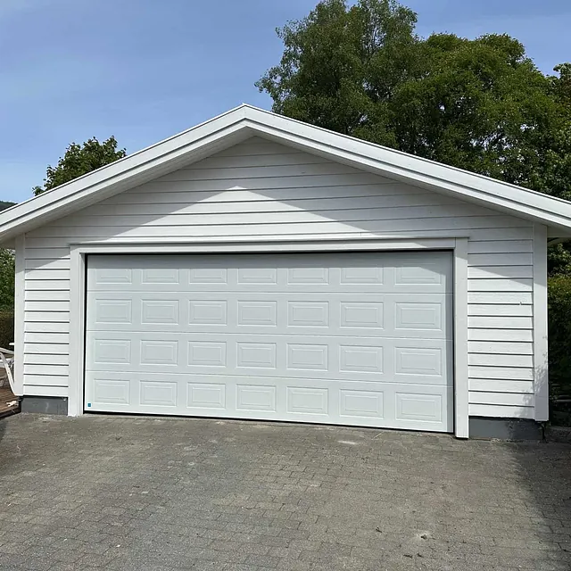 Hvit frittstående garasje med strukturert port og liggende kledning i hageområde.