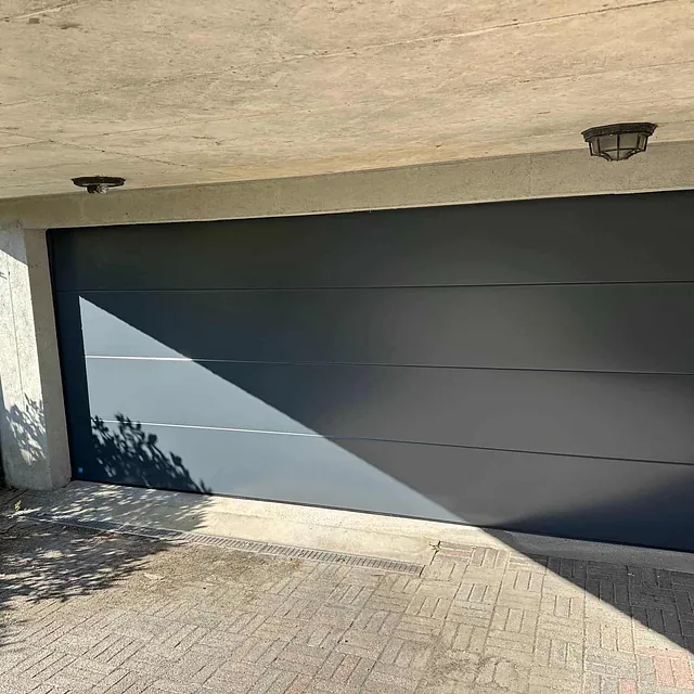 Garasje med svart port og betongtak, delvis i skygge fra nærliggende trær.