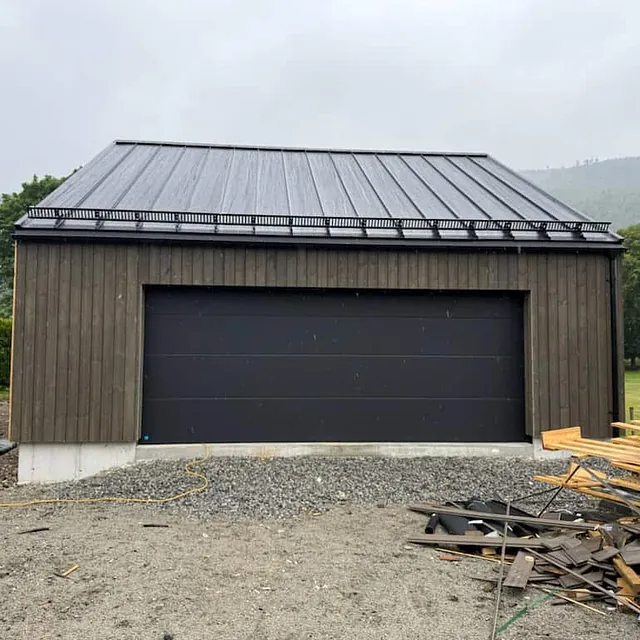 Frittstående garasje med svart port, stående trekledning og mørkt tak i regnvær.